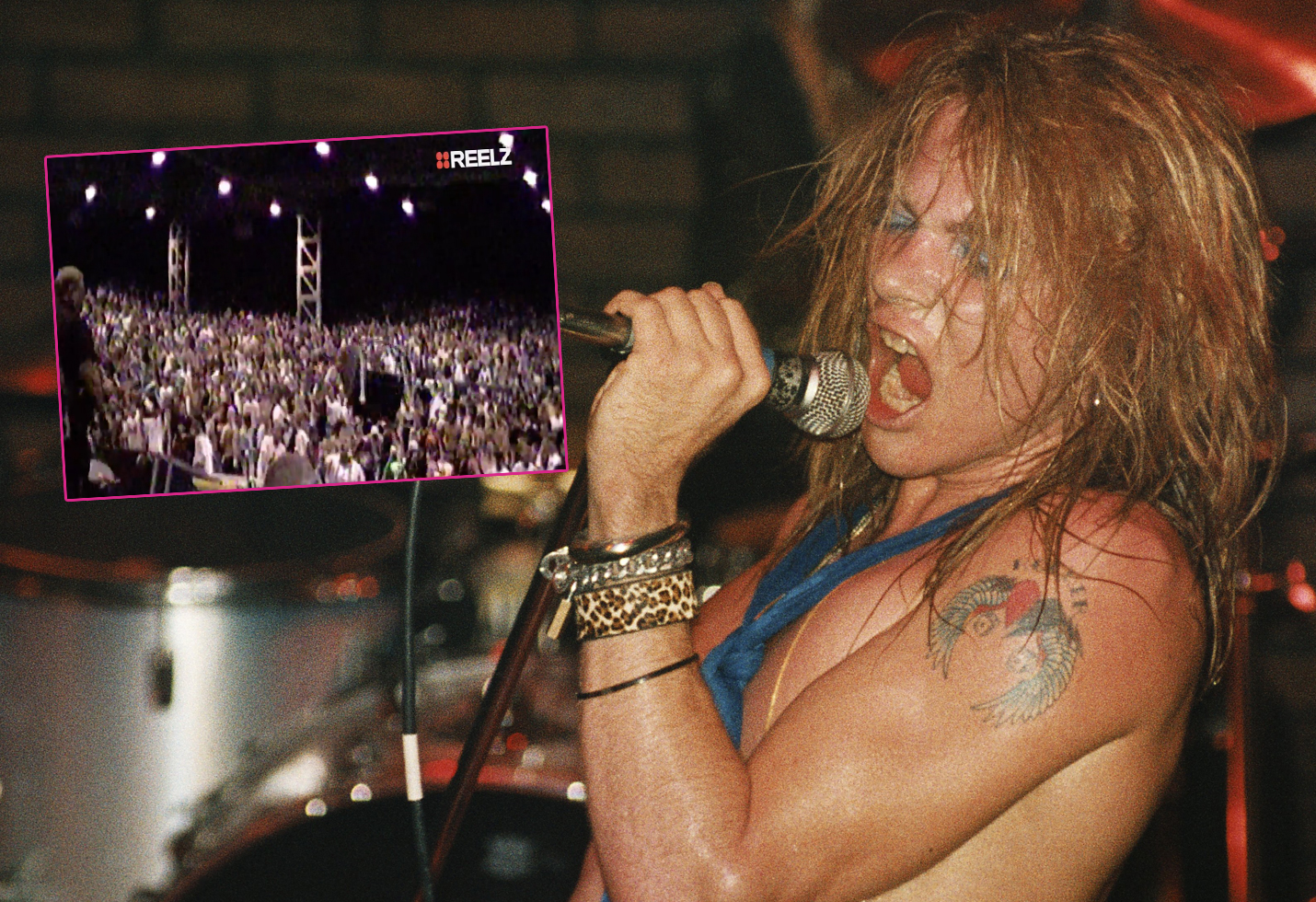 Axl Rose grips a microphone while wearing no shirt and sporting a blue scarf.