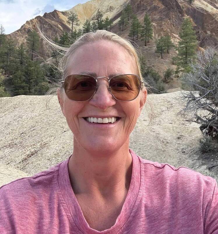 Christine Brown takes a selfie outdoors in front of rocky hills and pine trees, wearing sunglasses and a pink shirt.