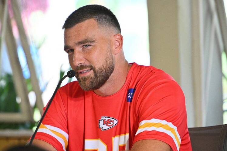 Travis Kelce speaks at a press conference wearing a red Kansas City Chiefs shirt with the team logo.